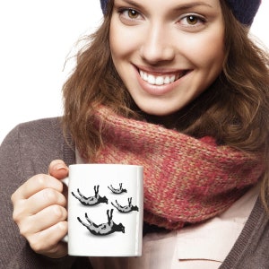 May include: White ceramic mug with a black and white graphic of four cows upside down. The mug is being held by a person wearing a pink scarf and a blue beanie.