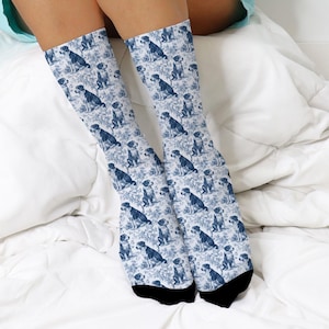 May include: Knee-high socks with a blue and white toile design, showcasing dog illustrations. The socks have black heels and toes. The dogs are shown in different poses, set against a backdrop of floral patterns.