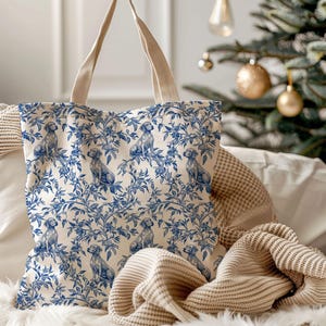 Puede incluir: Un bolso tote de lona con un estampado de perro y flores en azul y blanco. El bolso tiene asas beige y un estampado a juego en ambos lados. El bolso está sobre una superficie blanca con una manta beige y un árbol de Navidad al fondo.