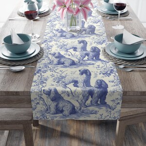 May include: A long rectangular table runner featuring a blue and white toile pattern with illustrations of spaniel dogs interspersed among floral branches. The fabric is laid flat across a wooden table set with blue bowls and wine glasses.