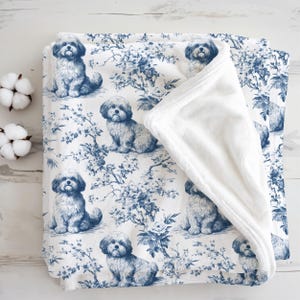 May include: A white and blue blanket with a repeating pattern of a dog breed and floral elements. The blanket has a soft, white, plush underside. Two cotton bolls are visible on the left side of the image.