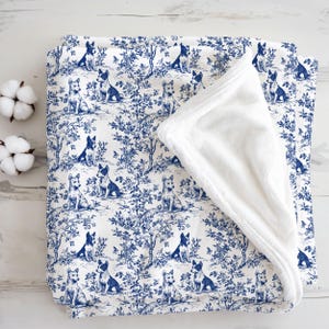 May include: A baby blanket with a blue and white toile pattern featuring dogs in a landscape scene. The blanket has a soft, white, plush underside. Two cotton bolls are visible on the left side of the image.