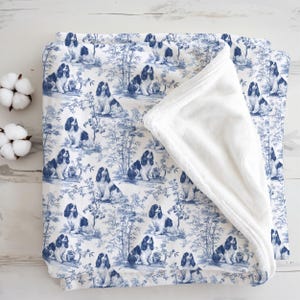 Peut inclure: Une couverture pliée avec un motif de toile de Jouy bleu et blanc représentant des Cavalier King Charles Spaniels et du bambou. La couverture a un dessous en peluche blanc et doux. Deux boules de coton sont visibles.