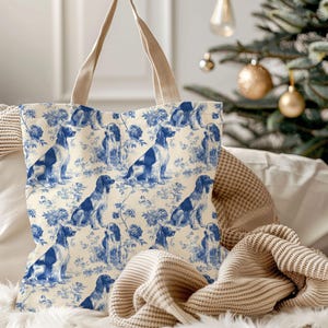 May include: A cream-coloured tote bag featuring a repeating blue toile pattern of seated spaniel dogs interspersed with floral motifs. The bag has two beige fabric handles and is constructed from a soft, woven material with a structured shape.