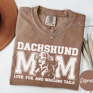 May include: Espresso-coloured t-shirt featuring the text "DACHSHUND MOM" in white block letters. Below the text is a graphic of a dachshund dog. The text "LOVE, FUR, AND WAGGING TAILS" is printed below the dog graphic. A silver necklace with a pendant is on the shirt.