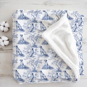 May include: A folded baby blanket with a blue and white toile pattern featuring Dalmatian dogs and trees. The blanket has a soft, white, plush underside. Two cotton buds are visible on the left side of the image.