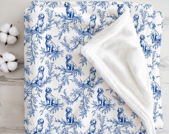 May include: A white blanket with a blue toile pattern featuring dogs and foliage. The blanket is folded, revealing a soft, white, plush interior. Two cotton bolls are visible in the upper left corner, adding a touch of natural texture.