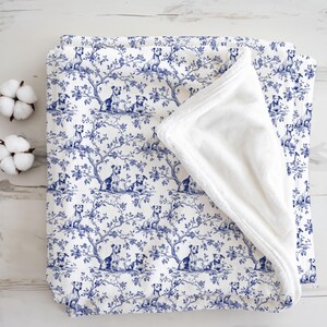 May include: A white and blue blanket with a dog and tree pattern. The blanket has a soft, white, plush interior. The design features dogs sitting under trees in a toile style. Two cotton buds are on the left.