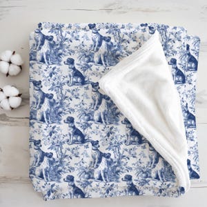 May include: A folded blanket with a blue and white toile pattern featuring dogs and floral elements. The blanket has a soft, white, plush underside. Two cotton bolls are visible in the upper left corner.