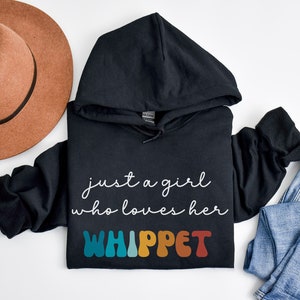 Może przedstawiać: Czarna bluza z kapturem z napisem "just a girl who loves her WHIPPET" w białym kolorze i słowem "WHIPPET" w kolorach tęczy.