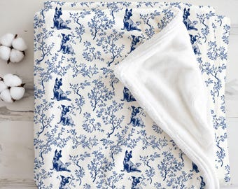 May include: A blanket with a white and blue dog and tree pattern. The blanket has a soft, white, plush underside. The design is repeated across the surface. Two cotton bolls are visible in the upper left corner.