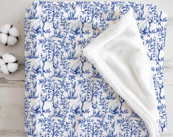 May include: A blue and white patterned blanket with a repeating design of dogs and foliage. The blanket has a soft, white, plush underside. The design is in a classic blue and white colour scheme. The blanket is folded over to show the contrasting textures.