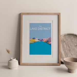 May include: A framed print of a colourful illustration of a lake in the Lake District, England. The print has a light blue background with the text "THE LAKE DISTRICT" in black.