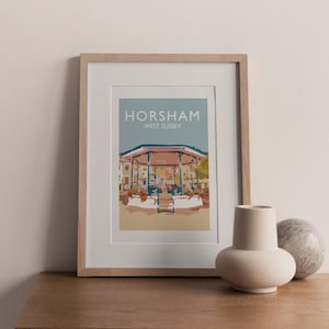 May include: A framed art print depicting the Horsham bandstand in West Sussex. The print features a light blue sky background with the word "Horsham" and "West Sussex" in white text. The bandstand is illustrated in beige, brown, and green.
