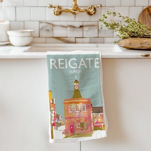 May include: A Reigate, Surrey, tea towel features a colorful illustration of a building with a clock tower. The towel has the word "REIGATE" in large white letters at the top, with "SURREY" below. The building is in shades of orange, pink, and yellow.