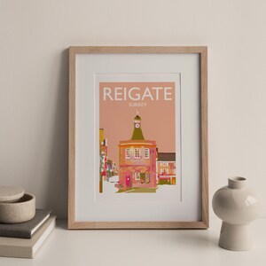 May include: Framed art print featuring a colorful illustration of Reigate, Surrey, with the town's name in white block letters. The artwork depicts a building with a clock tower and is set against a peach-colored background. The frame is light brown.