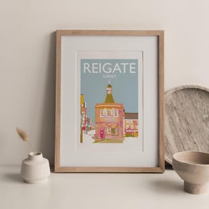 May include: A framed print of a colorful illustration of a building in Reigate, Surrey. The building has a clock tower and is surrounded by other buildings. The print is on a light blue background with the text "Reigate, Surrey" in white.