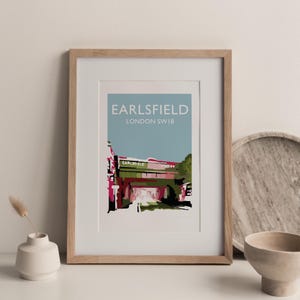 May include: Framed art print featuring a stylized illustration of Earlsfield Station in London, SW18. The artwork displays the station's architecture in shades of pink, green, and black against a light blue sky. The print is encased in a light wood frame.