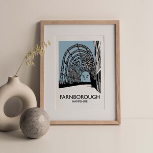 Puede incluir: Una impresión enmarcada de una ilustración en blanco y negro de una gran estructura metálica con arcos. La estructura está sobre un fondo azul claro. El texto "FARNBOROUGH HAMPSHIRE" está impreso debajo de la ilustración.