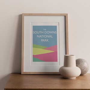May include: Framed print featuring the text "THE SOUTH DOWNS NATIONAL PARK" above a colorful landscape illustration. The artwork is in a light wood frame, displayed on a wooden surface, with a beige vase and a stone sphere.
