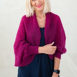 May include: A woman wearing a magenta knitted shrug with long sleeves over a dark blue dress. She is smiling and looking at the camera.