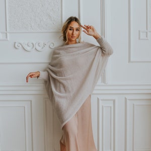 May include: A woman wearing a light beige oversized knit sweater with long sleeves and a long, light beige skirt. She is standing in front of a white wall with decorative molding.