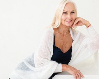 May include: A woman with blonde hair is wearing a white cardigan over a dark blue dress. She is sitting on a white staircase and smiling at the camera.