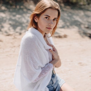 May include: A woman wearing a white knitted cardigan with a lace sleeve and denim shorts. She is standing on a sandy beach.