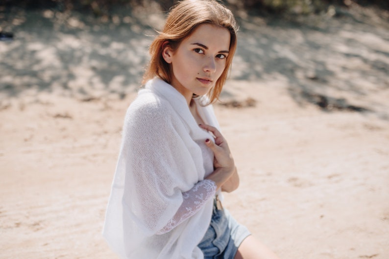 May include: A woman wearing a white knitted cardigan with a lace sleeve and denim shorts. She is standing on a sandy beach.