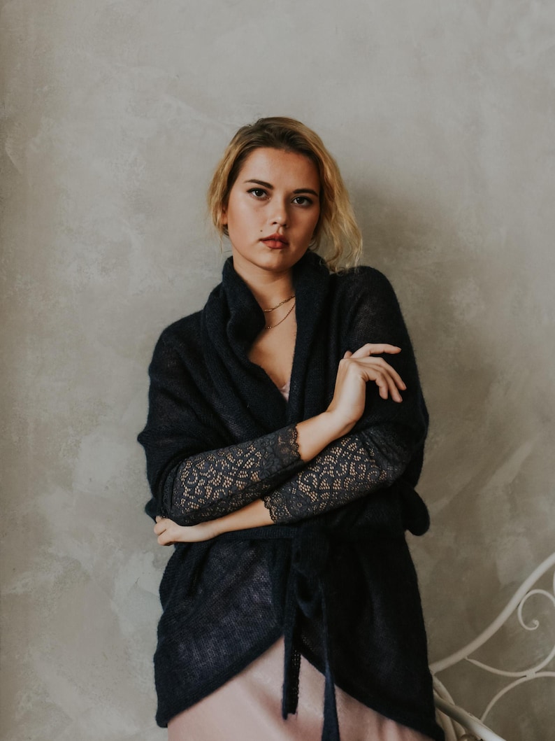 May include: A woman wearing a dark blue knitted cardigan with lace detailing on the sleeves. The cardigan is open in the front and tied at the waist. She is wearing a pink slip dress underneath.