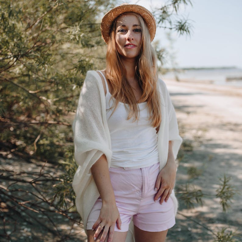 May include: A woman wearing a white cardigan, a white tank top, and pink shorts. She is wearing a straw hat and has long blonde hair.