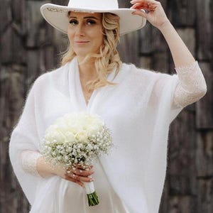 May include: A woman wearing a white knit shawl and a white hat with a beaded trim holds a bouquet of white roses and baby's breath.