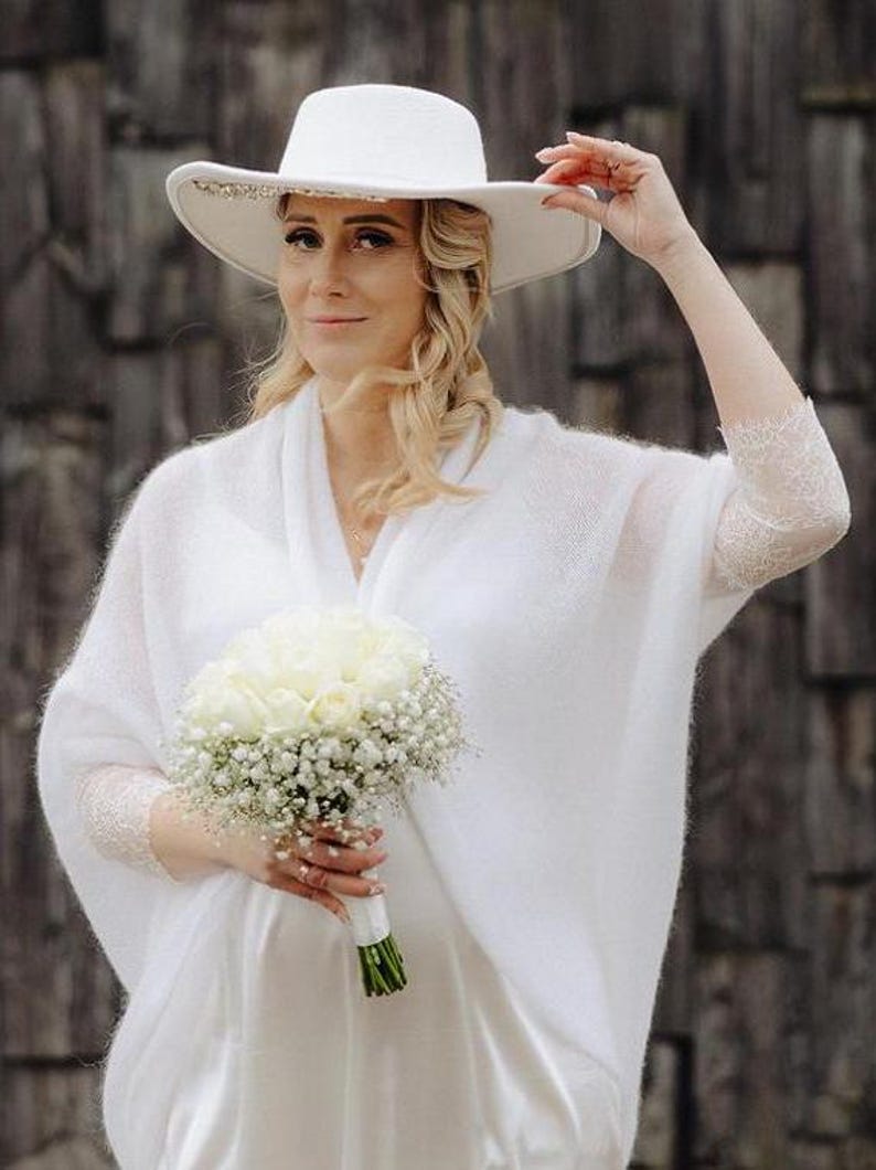 May include: A woman wearing a white knit shawl and a white hat with a beaded trim holds a bouquet of white roses and baby's breath.