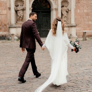 May include: A bride and groom walking away from the camera on a cobblestone street. The bride is wearing a white gown with a long veil and a floral bouquet. The groom is wearing a dark suit. They are walking in front of a building with an arched doorway.