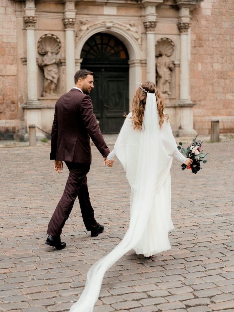 May include: A bride and groom walking away from the camera on a cobblestone street. The bride is wearing a white gown with a long veil and a floral bouquet. The groom is wearing a dark suit. They are walking in front of a building with an arched doorway.