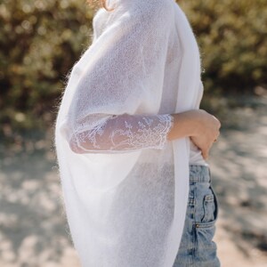 May include: A white knitted cardigan with lace sleeves. The cardigan is open at the front and has a relaxed fit. The model is wearing the cardigan over a white vest top and blue denim shorts.