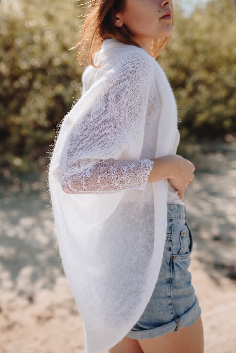 May include: A white knitted cardigan with lace sleeves. The cardigan is open at the front and has a relaxed fit. The model is wearing the cardigan over a white vest top and blue denim shorts.