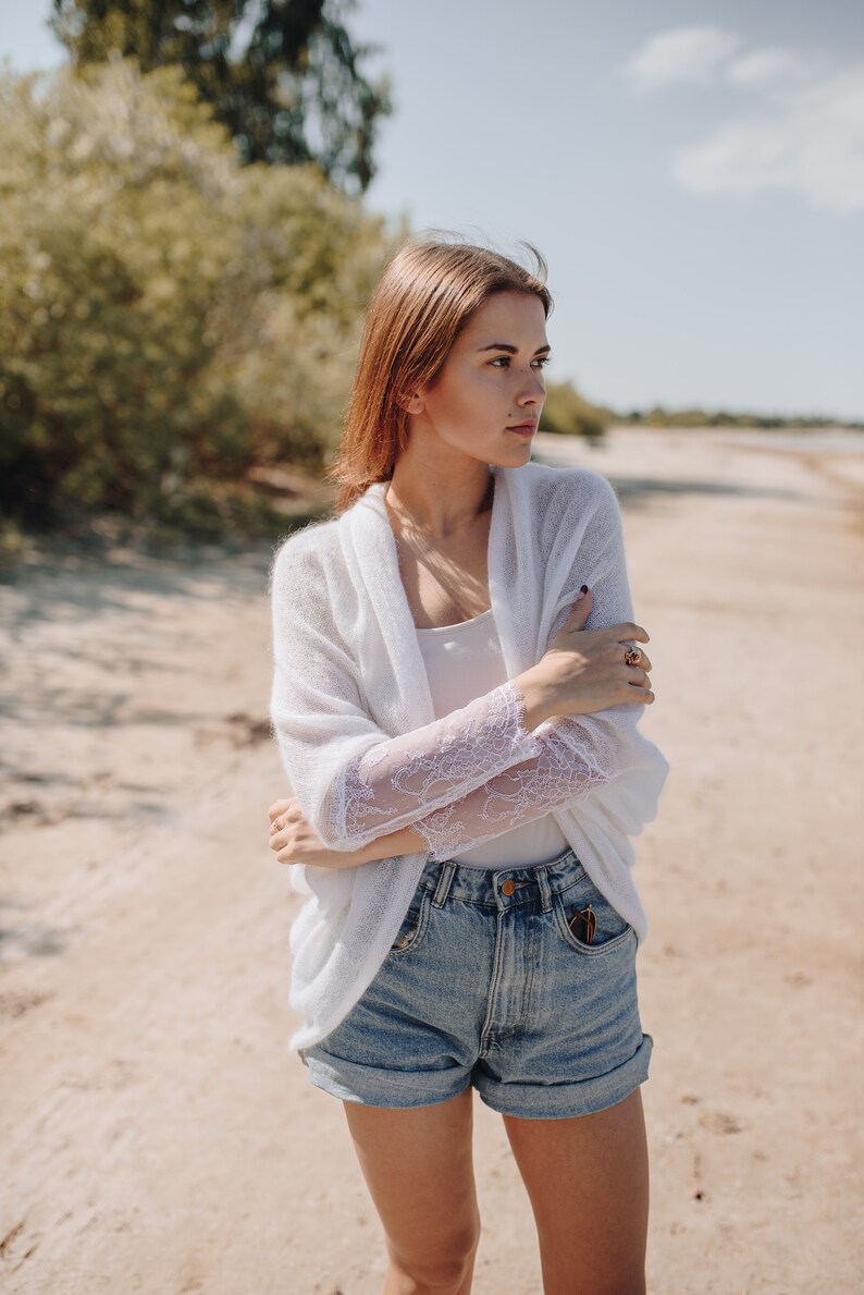 May include: A woman wearing a white cardigan with lace sleeves and denim shorts. She is standing on a sandy beach and looking off to the side.