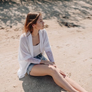 May include: A woman wearing a white cardigan with lace detailing sits on a sandy beach. She is wearing denim shorts and has her legs crossed.