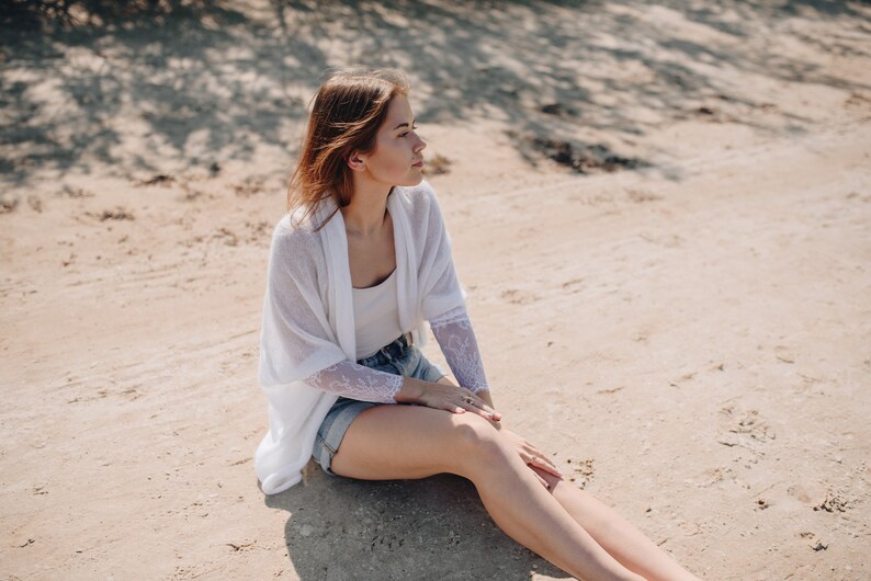 May include: A woman wearing a white cardigan with lace detailing sits on a sandy beach. She is wearing denim shorts and has her legs crossed.