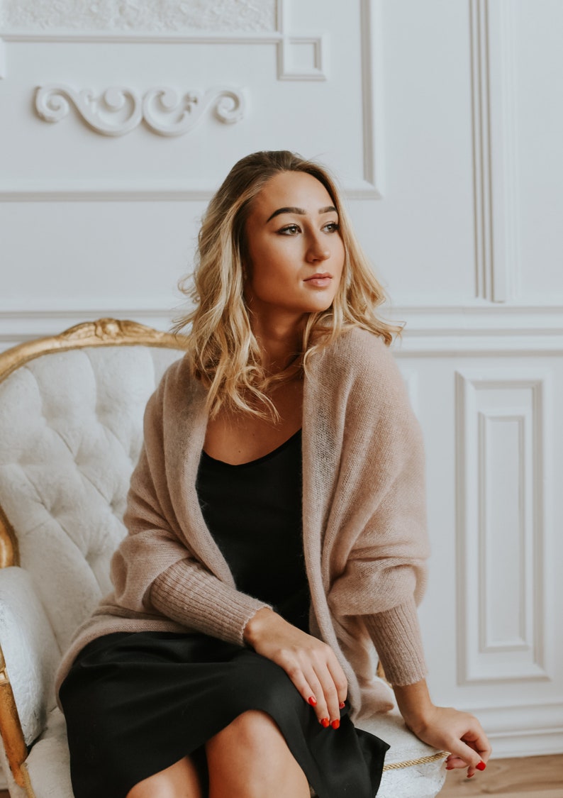 May include: A woman wearing a light brown knitted cardigan over a black dress. She is sitting in a white chair with gold accents.