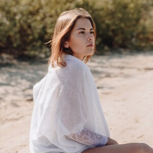 May include: A woman sits on a sandy beach wearing a white knit top with lace detailing and denim shorts. She is looking off to the side.