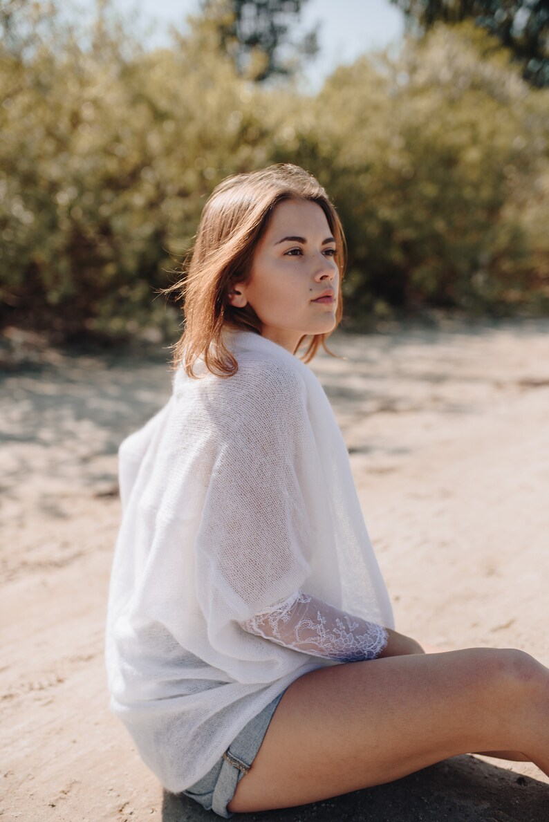 May include: A woman sits on a sandy beach wearing a white knit top with lace detailing and denim shorts. She is looking off to the side.