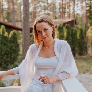 May include: A woman wearing a white knitted cardigan with lace trim, a white tank top, and white pants. She is standing in front of a white railing with a forest background.
