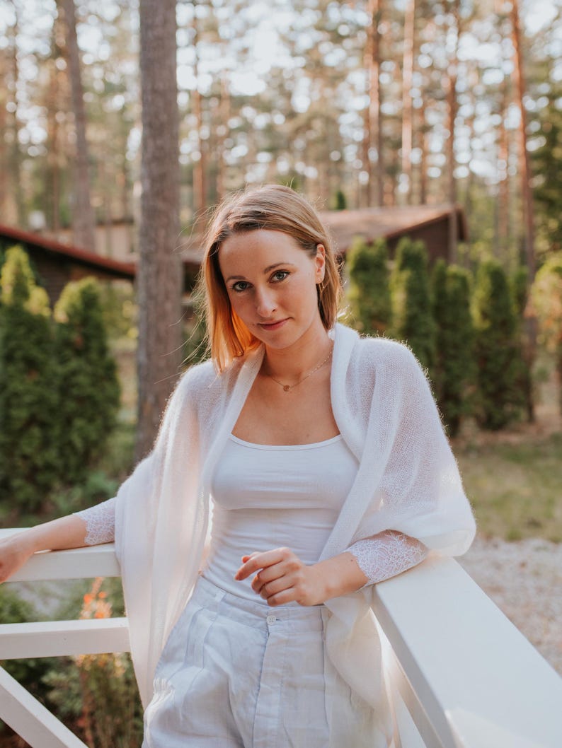 May include: A woman wearing a white knitted cardigan with lace trim, a white tank top, and white pants. She is standing in front of a white railing with a forest background.