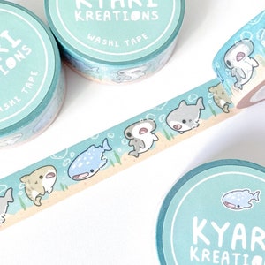 A row of cute shark illustrations printed on a roll of washi tape. Shark species including: hammerheads, great whites, tiger sharks, and whale sharks.  3 additional rolls of washi tapes lying around in background for display purposes