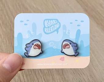May include: A pair of blue and white shark earrings on a card with the text "Kyar! Kreations" and the website address "www.kyarikreations.com".