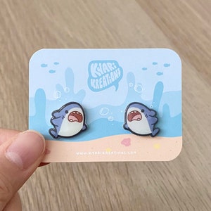 May include: A pair of blue and white shark earrings on a card with the text "Kyar! Kreations" and the website address "www.kyarikreations.com".