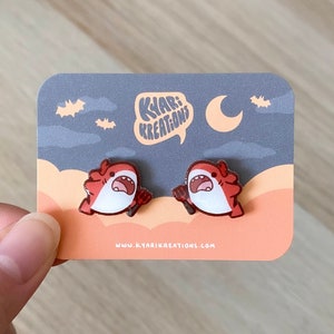 Puede incluir: Un par de pendientes de tiburón rojo y blanco en una tarjeta con fondo de cielo nocturno. La tarjeta dice "Kyar! Kreations" y "www.kyarikreations.com".