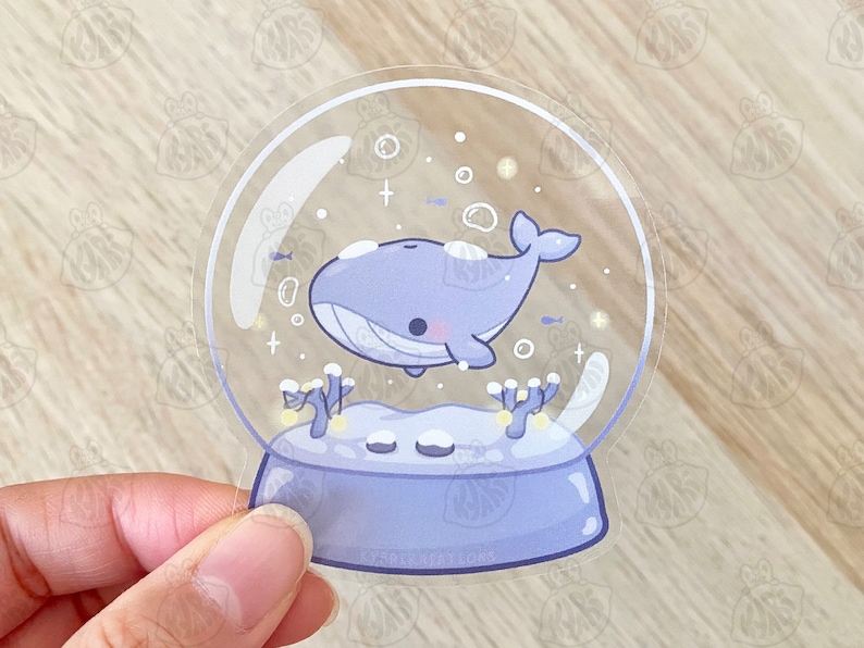 Pode incluir: Um globo de neve de vidro transparente com uma baleia azul dentro. A baleia est&aacute; nadando em uma cena nevada branca com pequenos peixes azuis e estrelas brancas. O globo de neve est&aacute; em uma base azul claro.